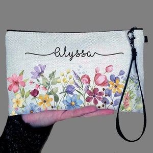 May include: A white canvas zippered pouch with a black strap. The pouch has a floral design with pink, yellow, blue, and purple flowers. The name "Alyssa" is written in black cursive on the front of the pouch.