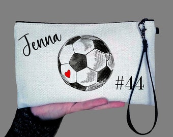 Op de afbeelding: Witte canvas toilettas met een zwart-wit voetbalontwerp, de naam "Jenna" en het nummer "#44" erop gedrukt. De tas heeft een zwarte rits en een zwarte band.