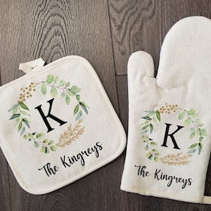 May include: A set of two kitchen pot holders with a green and gold floral design and the letter "K" in the center. The pot holders also have the text "The Kingreys" printed on them.