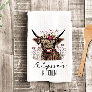 May include: A white kitchen towel with a brown and white Highland cow wearing a floral crown. The text "Alyssa's Kitchen" is printed below the cow.
