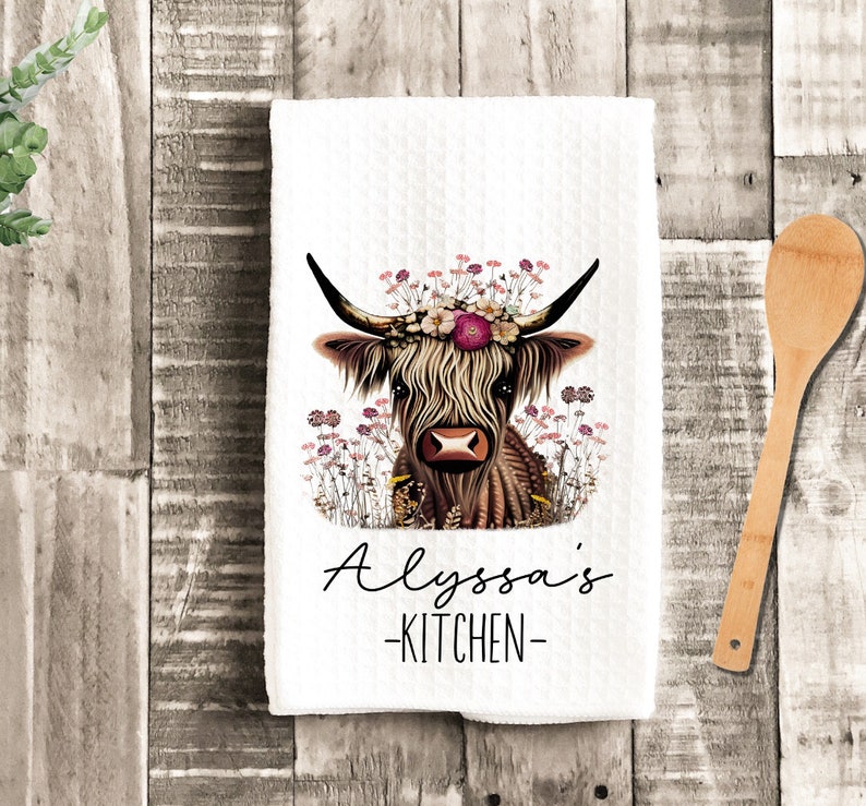 May include: A white kitchen towel with a brown and white Highland cow wearing a floral crown. The text "Alyssa's Kitchen" is printed below the cow.