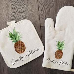 May include: A set of two kitchen accessories featuring a pineapple design. The set includes a pot holder and an oven mitt. Both items are made of a natural linen fabric and feature the text "Caitlyn's Kitchen" in black cursive writing.