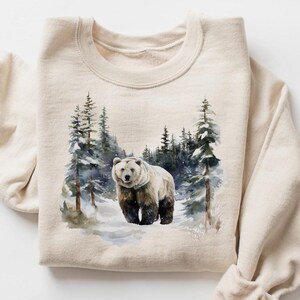 May include: Beige sweatshirt featuring a watercolor painting of a bear in a snowy forest. The bear is brown and white, standing in the snow with evergreen trees in the background. The sweatshirt has a crew neck and long sleeves.