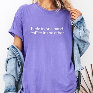 Pu&ograve; includere: T-shirt lilla con la scritta bianca "bible in one hand, coffee in the other". La maglietta ha le maniche arrotolate e una vestibilit&agrave; rilassata. Una giacca di jeans azzurra &egrave; indossata sulle spalle.