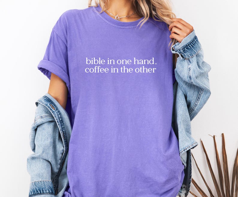 Pu&ograve; includere: T-shirt lilla con la scritta bianca "bible in one hand, coffee in the other". La maglietta ha le maniche arrotolate e una vestibilit&agrave; rilassata. Una giacca di jeans azzurra &egrave; indossata sulle spalle.