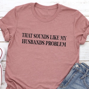 May include: A dusty rose colored t-shirt with the text "THAT SOUNDS LIKE MY HUSBANDS PROBLEM" printed in black.