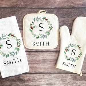 May include: A set of three kitchen items featuring a wreath design with a green and red color scheme. The items include a potholder, a tea towel, and an oven mitt. Each item has the letter "S" and the word "SMITH" printed on it.