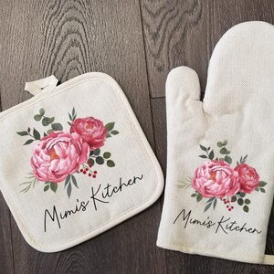 May include: A set of two kitchen pot holders with a floral design. The pot holders are beige with pink flowers and green leaves. The text "Mimi's Kitchen" is printed on both pot holders.