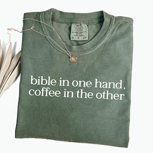 Pu&ograve; includere: Una maglietta verde oliva con la scritta bianca "bible in one hand, coffee in the other". Una collana color oro con un ciondolo &egrave; drappeggiata sulla maglietta. La maglietta &egrave; piegata ordinatamente su una superficie bianca.
