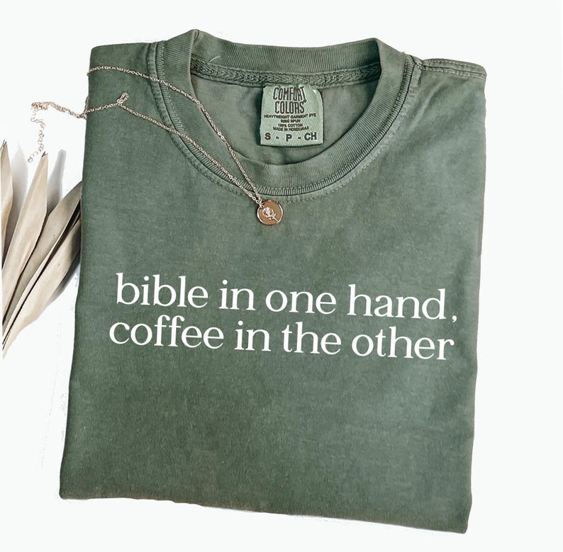 Pu&ograve; includere: Una maglietta verde oliva con la scritta bianca "bible in one hand, coffee in the other". Una collana color oro con un ciondolo &egrave; drappeggiata sulla maglietta. La maglietta &egrave; piegata ordinatamente su una superficie bianca.