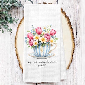 May include: White waffle-weave kitchen towel featuring a floral teacup design. The teacup is blue and white striped, filled with pink tulips, white and yellow flowers, and blue accents. Text reads "my cup runneth over" and "psalm 23:5".