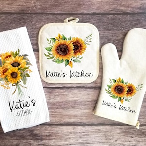 May include: A set of three kitchen towels featuring a sunflower design and the text "Katie's Kitchen". The set includes a potholder, a tea towel, and an oven mitt.