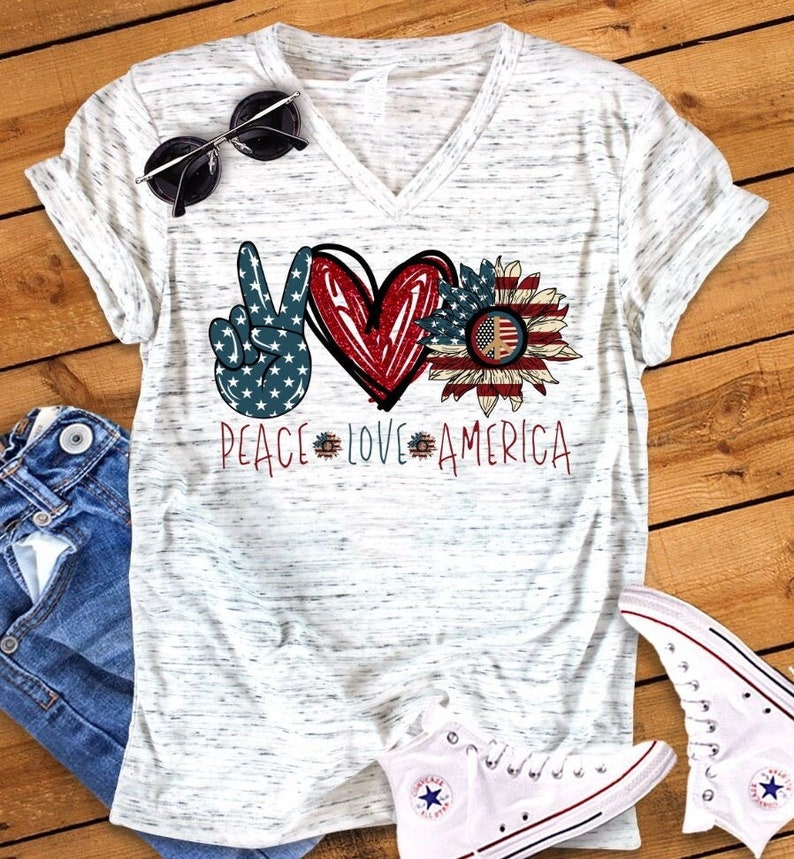 May include: White t-shirt with a peace sign, heart, and sunflower design. The design is in red, white, and blue with a peace sign in the center of the sunflower. The text "Peace Love America" is printed below the design.