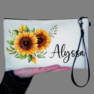 May include: White canvas zippered pouch with a black strap. The pouch features a watercolor design of sunflowers and green leaves. The name "Alyssa" is printed in black cursive lettering.