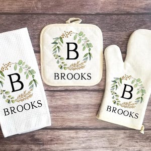 May include: A set of three kitchen towels with a green and gold floral wreath design. The towels feature the letter "B" in the center of the wreath and the name "BROOKS" printed below.