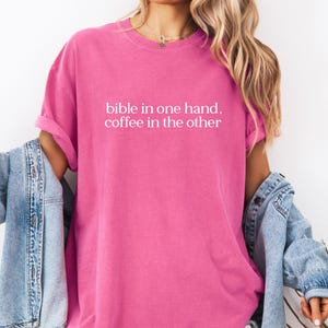 Pu&ograve; includere: Una maglietta rosa acceso con la scritta bianca "bible in one hand, coffee in the other". La maglietta ha maniche corte e una vestibilit&agrave; rilassata. Una giacca di jeans azzurra &egrave; indossata sulle spalle.