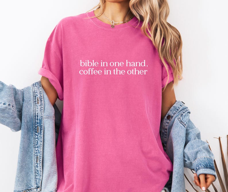 Pu&ograve; includere: Una maglietta rosa acceso con la scritta bianca "bible in one hand, coffee in the other". La maglietta ha maniche corte e una vestibilit&agrave; rilassata. Una giacca di jeans azzurra &egrave; indossata sulle spalle.