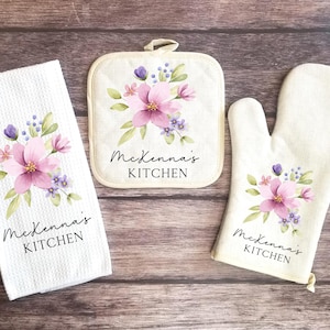 May include: A kitchen set featuring a white waffle-weave towel, a square pot holder, and an oven mitt. Each item is decorated with a floral design in pink, purple, and green, and the text "McKenna's KITCHEN".