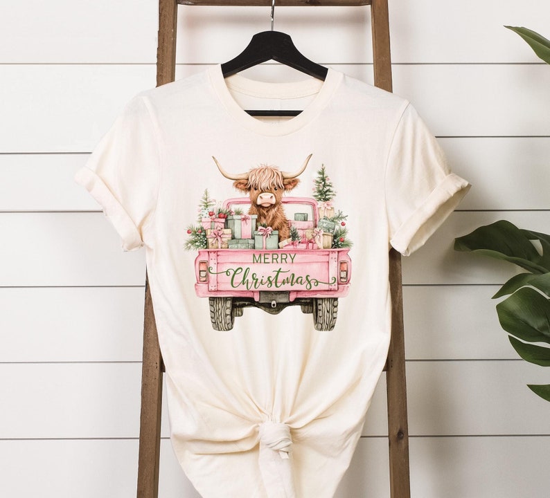 May include: Cream-colored t-shirt featuring a watercolor illustration of a Highland cow in a pink pickup truck filled with gifts and Christmas trees. The text "MERRY Christmas" is written in a script font on the truck's door. The shirt is hanging on a black hanger.