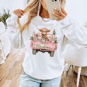 May include: White sweatshirt featuring a pink pickup truck graphic with a Highland cow, Christmas presents, and text that reads "Merry Christmas." The truck bed is filled with gifts and greenery.