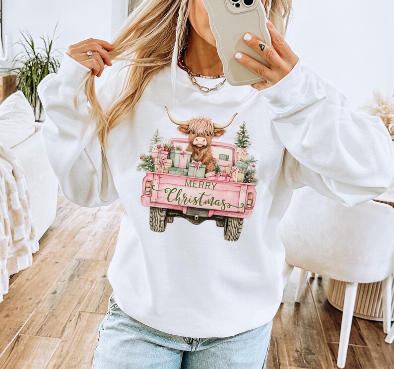 May include: White sweatshirt featuring a pink pickup truck graphic with a Highland cow, Christmas presents, and text that reads "Merry Christmas." The truck bed is filled with gifts and greenery.