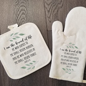 May include: A set of two white cotton kitchen accessories with a green leafy design and a bible verse. The pot holder and oven mitt feature the text "I am the bread of life. He who comes to me shall never hunger, and he who believes in me shall never thirst. John 6:35".