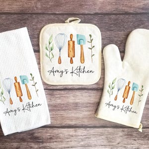 May include: A set of three kitchen towels with a white background and a design of kitchen utensils and green leaves. The towels have the text "Amy's Kitchen" printed on them.