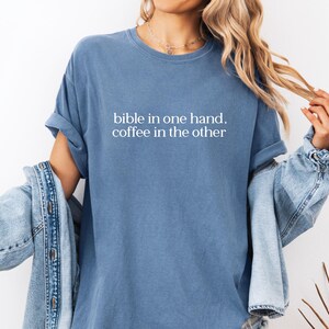 Pu&ograve; includere: Una maglietta blu con la scritta bianca "bible in one hand, coffee in the other". Le maniche sono arrotolate. Una giacca di jeans &egrave; indossata sopra la maglietta. Lo stile &egrave; casual e confortevole.