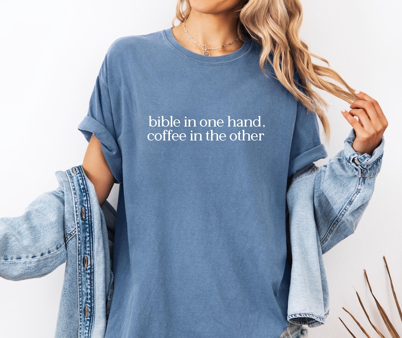 Pu&ograve; includere: Una maglietta blu con la scritta bianca "bible in one hand, coffee in the other". Le maniche sono arrotolate. Una giacca di jeans &egrave; indossata sopra la maglietta. Lo stile &egrave; casual e confortevole.