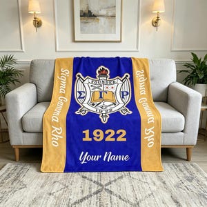 May include: A blue and gold blanket featuring the Sigma Gamma Rho crest, the founding year 1922, and space for a name. The blanket is draped over a grey sofa, with the text "Sigma Gamma Rho" in gold.
