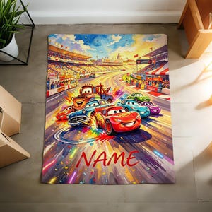 Pu&ograve; includere: Una coperta rettangolare con una vivace illustrazione pittorica di una pista da corsa con diverse auto antropomorfe, tra cui un'auto da corsa rossa e un carro attrezzi marrone, sotto un cielo luminoso. La parola NAME &egrave; stampata in rosso in basso.