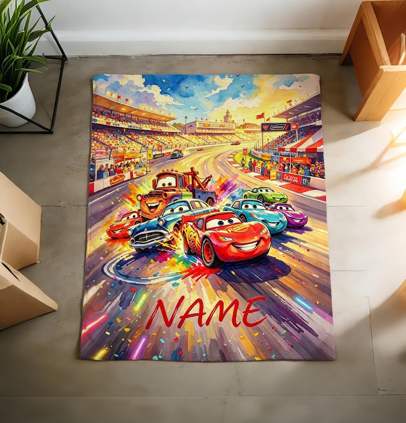 Pu&ograve; includere: Una coperta rettangolare con una vivace illustrazione pittorica di una pista da corsa con diverse auto antropomorfe, tra cui un'auto da corsa rossa e un carro attrezzi marrone, sotto un cielo luminoso. La parola NAME &egrave; stampata in rosso in basso.