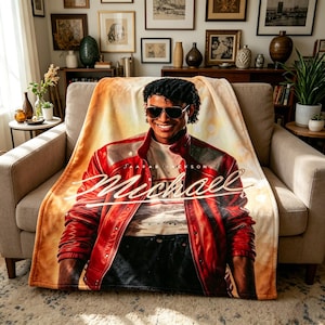 May include: A soft throw blanket displaying a portrait of Michael Jackson in a red leather jacket and sunglasses. The blanket features the name "Michael" in a stylish script. The background has a warm, sunset-like gradient.