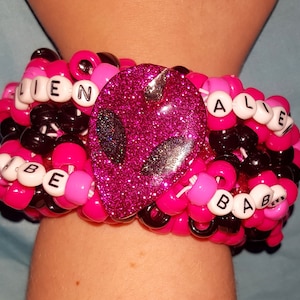 May include: A beaded bracelet with a pink glitter alien head in the center. The bracelet is made of pink, black, and white beads. The words "ALIEN", "ALIVE", and "VIBE BABE" are spelled out with white letter beads.
