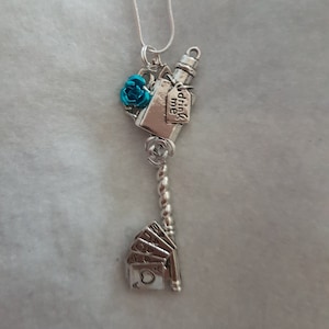 Peut inclure: Un collier pendentif argenté avec une breloque en forme de clé. La breloque comprend une petite rose bleue, une bouteille avec une étiquette portant l'inscription "drink me" et un éventail de cartes à jouer à la base, suspendu à une fine chaîne.