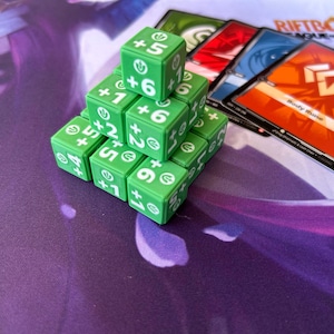Pode incluir: Uma pirâmide de dados verdes com números brancos e sinais de mais, ao lado de uma pilha de cartas de jogo coloridas. Os dados são marcados com números como +1, +2, +5 e +6. As cartas têm vários designs e texto, incluindo "Body Rune".