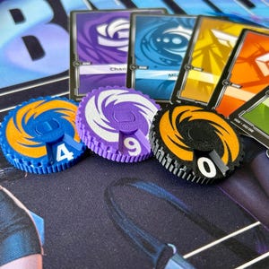 May include: Image of game components, including circular tokens with numbers and trading cards. The tokens are blue, purple, and orange, with white numbers. The cards have different designs and colours. The tokens are likely used for board games.