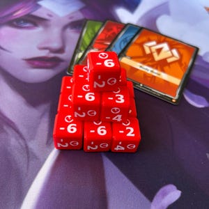 Pode incluir: Uma pirâmide de dados vermelhos com números e símbolos brancos, ao lado de cartas de jogo. Os dados estão dispostos em forma de pirâmide, com os números -6, -3, -4 e -2 visíveis. As cartas de jogo têm um design colorido.