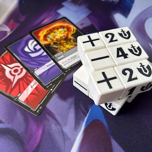 Pode incluir: Um dado de jogo branco em forma de cubo com símbolos e números pretos, incluindo mais, menos e os números 1, 2 e 4. Três cartas de jogo são visíveis, com texto como "Fury", "Gold" e "Chaos".