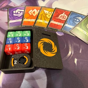 Pode incluir: Uma caixa preta contendo dados e cartas de jogo. Os dados são verdes, azuis e vermelhos, com números de -3 a +3. As cartas têm vários desenhos e texto. Uma carta preta com um desenho em espiral laranja também está presente.