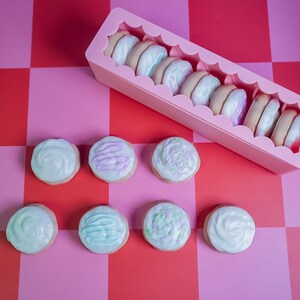 Pode incluir: Um recipiente retangular rosa cheio de macarons coloridos e v&aacute;rios biscoitos com cobertura em uma superf&iacute;cie xadrez rosa e vermelha. Os macarons t&ecirc;m v&aacute;rios recheios past&eacute;is e os biscoitos s&atilde;o decorados com redemoinhos e granulados.