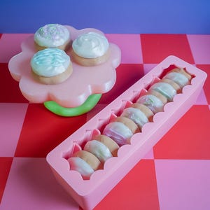 Pode incluir: Uma bandeja rosa em forma de flor cont&eacute;m tr&ecirc;s pequenos bolos com cobertura. Um recipiente retangular rosa cont&eacute;m v&aacute;rios biscoitos com cobertura. Os bolos e biscoitos t&ecirc;m cobertura em tons pastel. O fundo &eacute; um padr&atilde;o xadrez vermelho e rosa.