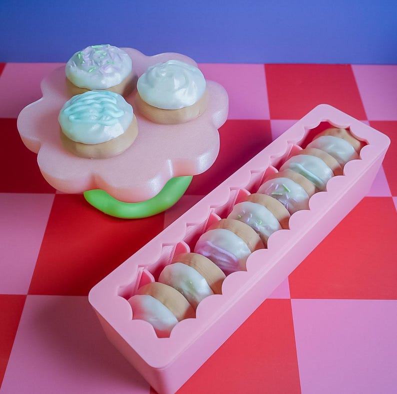 Pode incluir: Uma bandeja rosa em forma de flor cont&eacute;m tr&ecirc;s pequenos bolos com cobertura. Um recipiente retangular rosa cont&eacute;m v&aacute;rios biscoitos com cobertura. Os bolos e biscoitos t&ecirc;m cobertura em tons pastel. O fundo &eacute; um padr&atilde;o xadrez vermelho e rosa.
