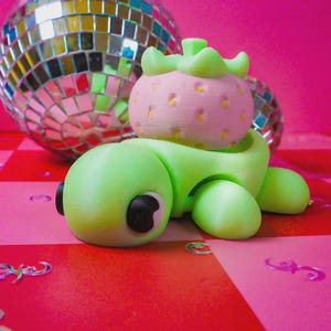 May include: A whimsical, 3D-printed sculpture of a strawberry atop a green, rounded body with black accents. The strawberry is pink with yellow speckles and green leaves. A disco ball is in the background, set against a pink and red checkered surface.