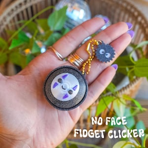 May include: A black glittery circular fidget clicker featuring a white face with purple accents and a small black soot sprite charm attached by a gold ball chain. The text NO FACE FIDGET CLICKER is printed in white at the bottom of the image.