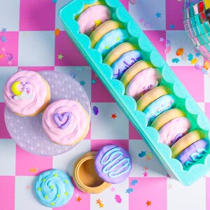 Pode incluir: Uma vista superior de biscoitos decorados e um recipiente de biscoitos em uma superf&iacute;cie xadrez rosa e branca. Os biscoitos t&ecirc;m cobertura pastel e v&aacute;rios desenhos, incluindo estrelas, luas e cora&ccedil;&otilde;es. Uma bola de discoteca est&aacute; no fundo.