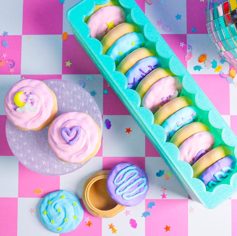 Pode incluir: Uma vista superior de biscoitos decorados e um recipiente de biscoitos em uma superf&iacute;cie xadrez rosa e branca. Os biscoitos t&ecirc;m cobertura pastel e v&aacute;rios desenhos, incluindo estrelas, luas e cora&ccedil;&otilde;es. Uma bola de discoteca est&aacute; no fundo.