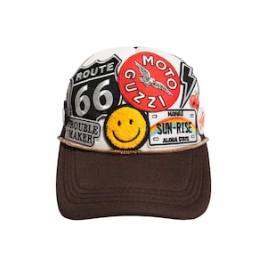 May include: A brown baseball cap with a white top covered in patches. Patches include a Route 66 sign, a smiley face, and a Moto Guzzi logo. The brim is brown, and the cap has a gold-colored trim.