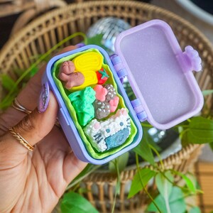 Puede incluir: Una caja bento de pl&aacute;stico morado en miniatura con tapa abatible y un revestimiento interior de color verde lima. La caja contiene peque&ntilde;os alimentos esculpidos, como una tortilla enrollada amarilla, un tomate rojo, br&oacute;coli y una bola de arroz.