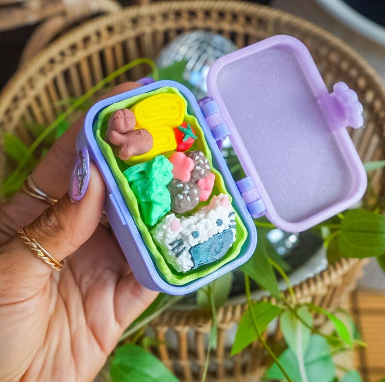 Puede incluir: Una caja bento de pl&aacute;stico morado en miniatura con tapa abatible y un revestimiento interior de color verde lima. La caja contiene peque&ntilde;os alimentos esculpidos, como una tortilla enrollada amarilla, un tomate rojo, br&oacute;coli y una bola de arroz.